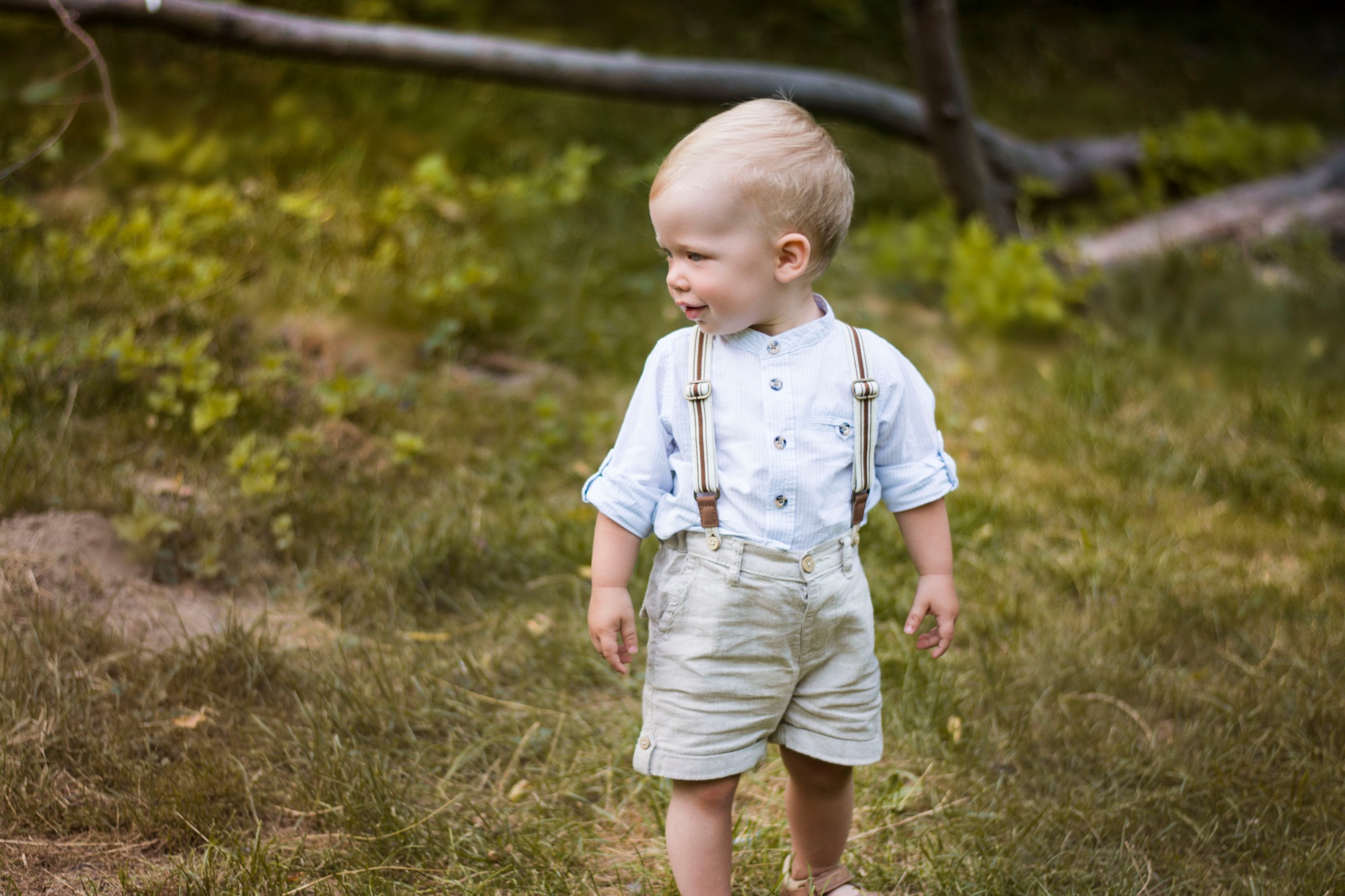 Mały chłopiec z blond włosami, ubrany w błękitną koszulę i lniane szorty z szelkami, stoi na trawie i patrzy w bok, na tle rozmytego krajobrazu.