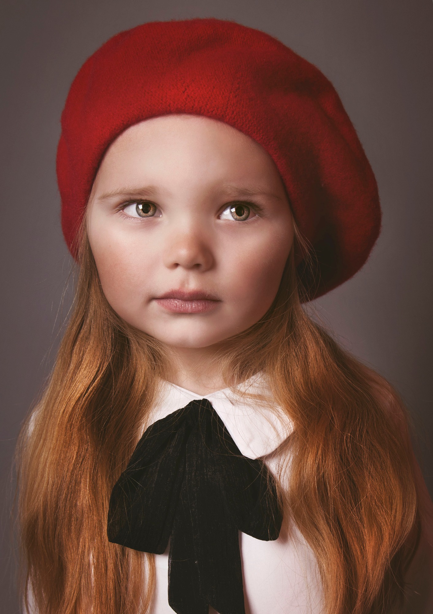 Portret dziewczynki z rudymi włosami i czerwonym berecie, ubrana w białą bluzkę z czarną kokardą, na szarym tle. Delikatne światło podkreśla rysy twarzy.