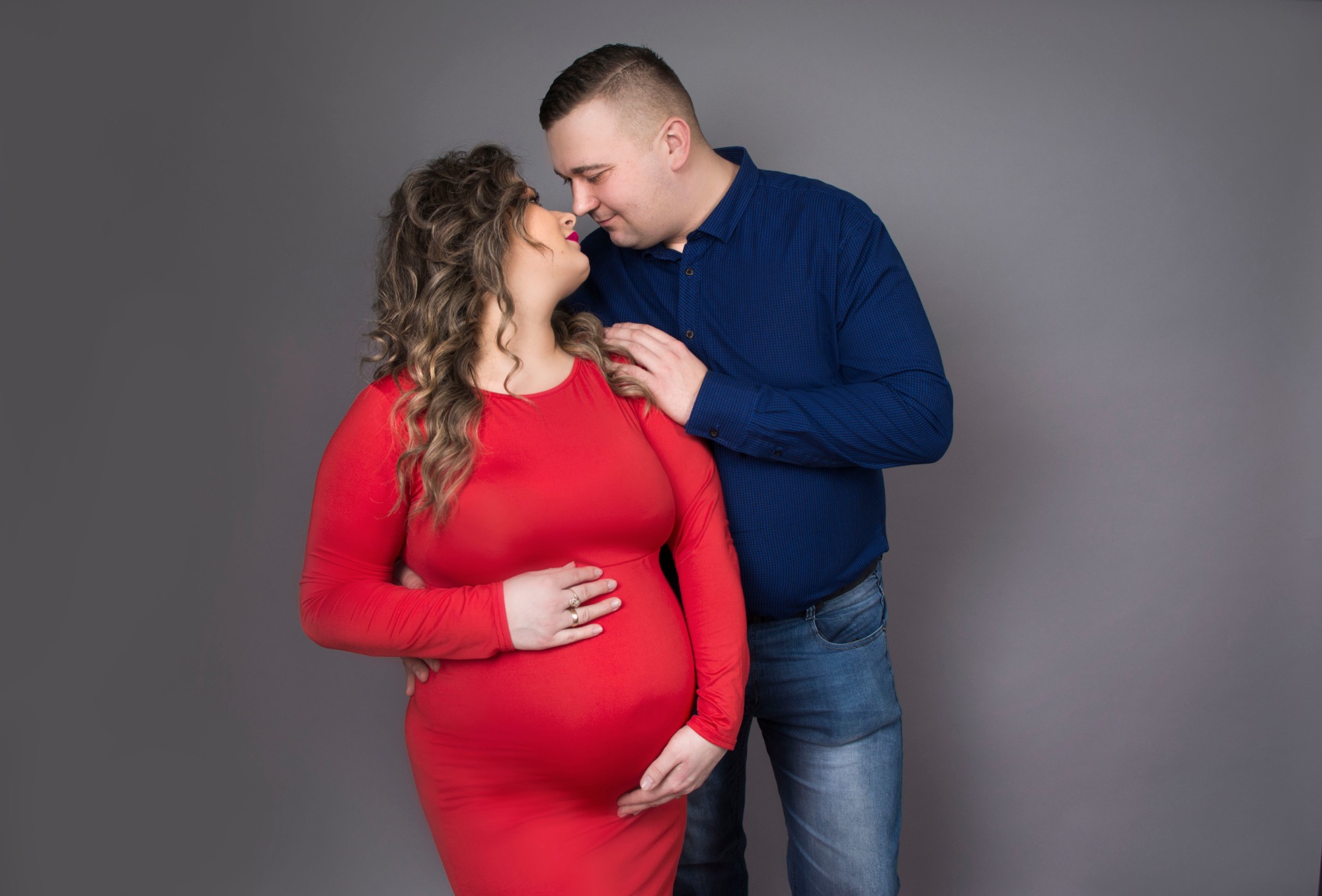 Portret przyszłych rodziców: kobieta w czerwonej sukience i mężczyzna w niebieskiej koszuli, oboje patrzą na siebie z czułością na tle szarego studia.