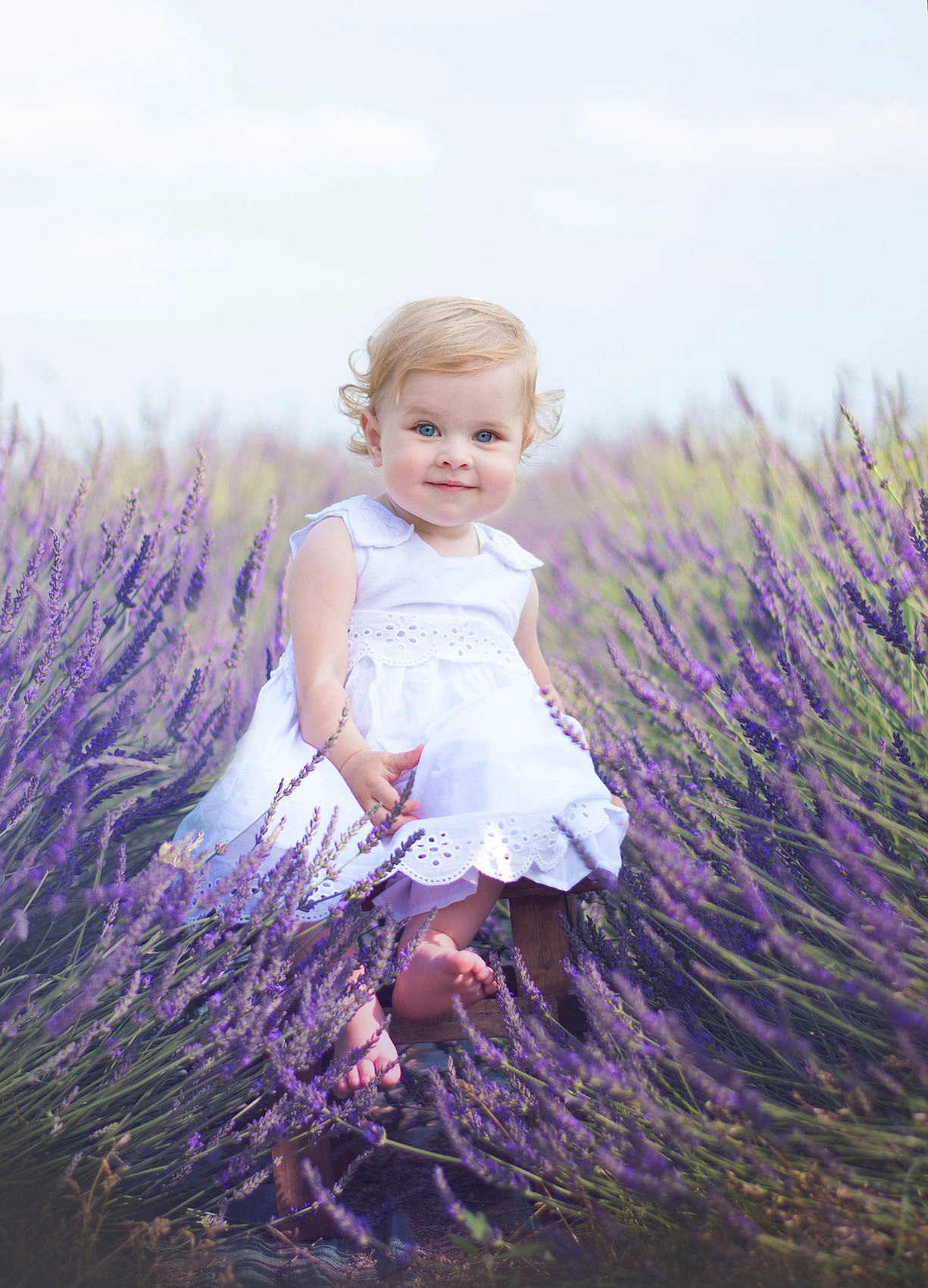 Uśmiechnięta dziewczynka w białej sukience siedzi na drewnianym stołku pośród kwitnącej lawendy, jej bose stopy delikatnie dotykają podłoża.