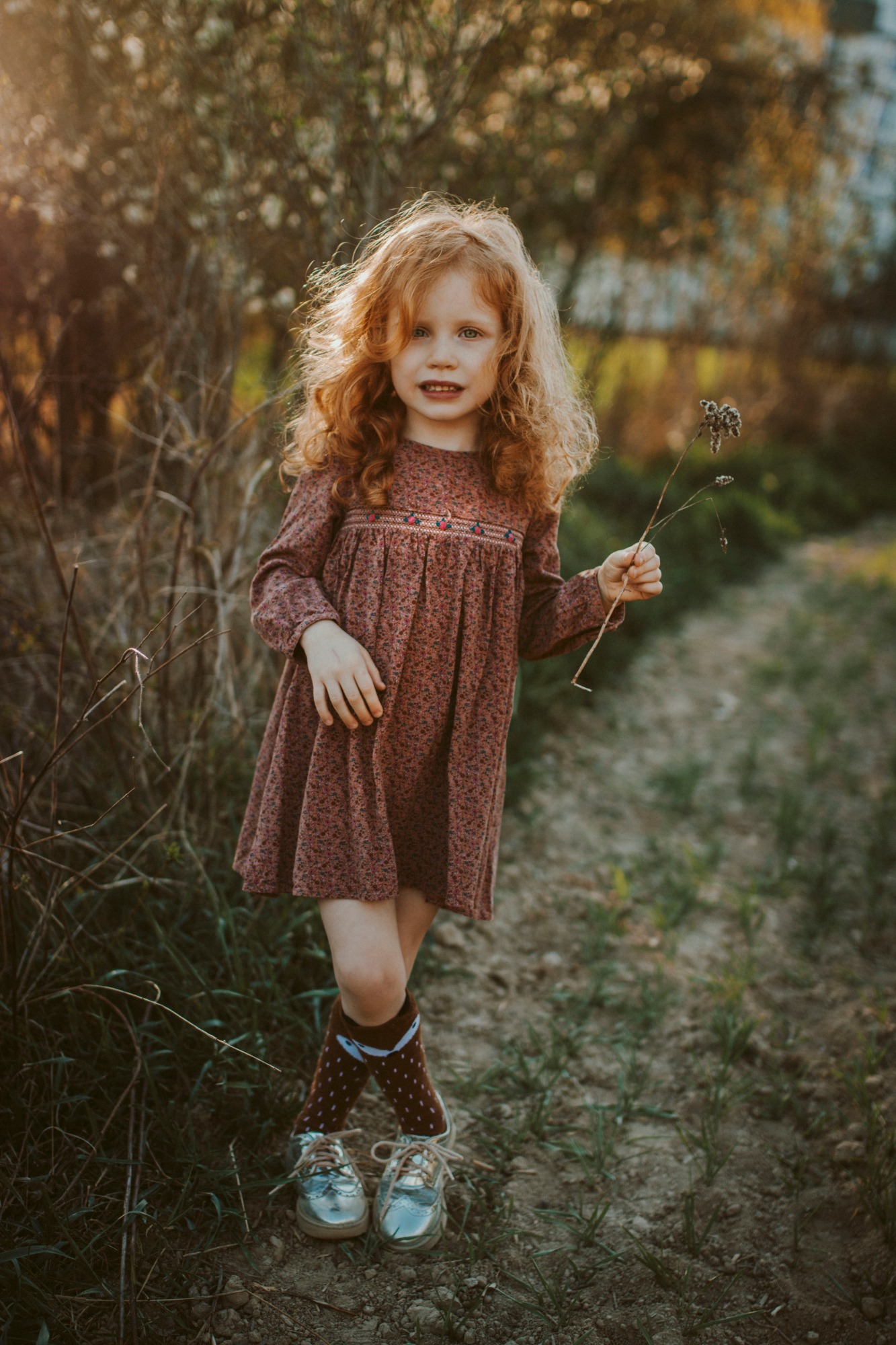 Ruda dziewczynka w kwiecistej sukience i srebrnych butach stoi na polnej drodze, trzymając suchą gałązkę, z rozczochranymi włosami i uśmiechem na twarzy.