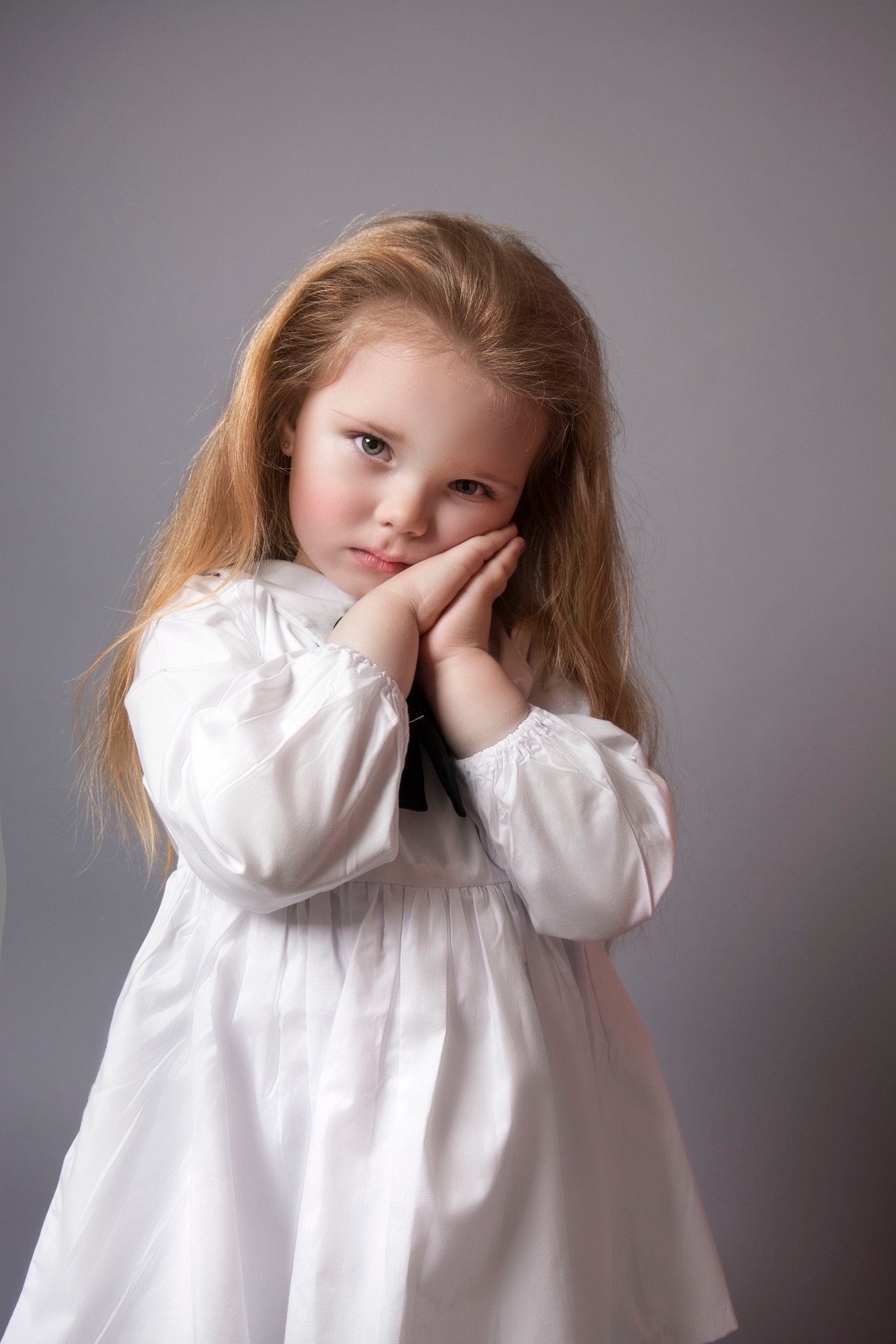 Portret małej dziewczynki z długimi blond włosami w białej sukience z czarną kokardką, opierającej twarz na dłoniach na szarym tle.
