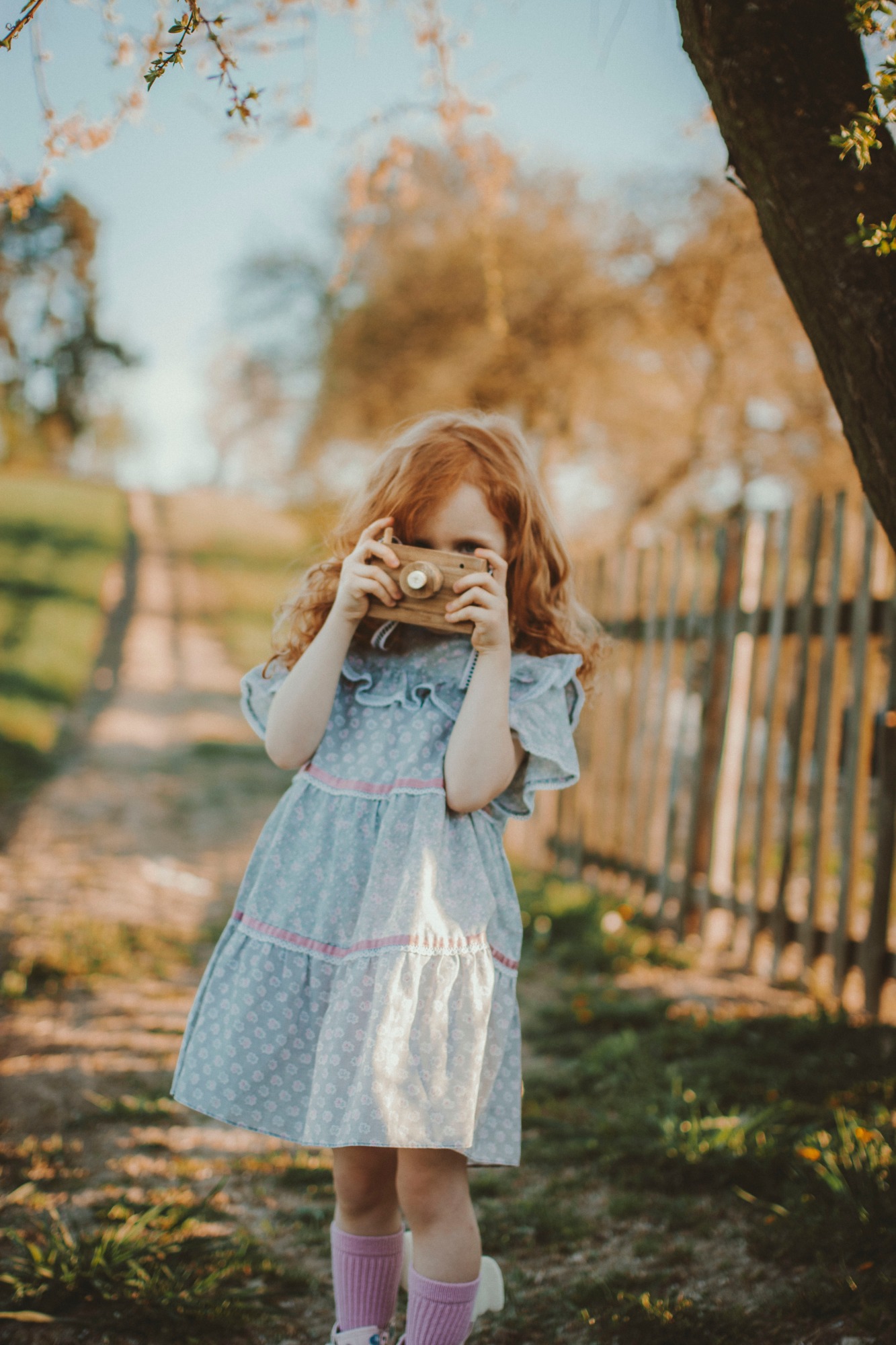 Dziewczynka z rudymi włosami w kwiecistej sukience trzyma drewniany aparat fotograficzny, pozując na tle wiejskiej ścieżki i drewnianego płotu.