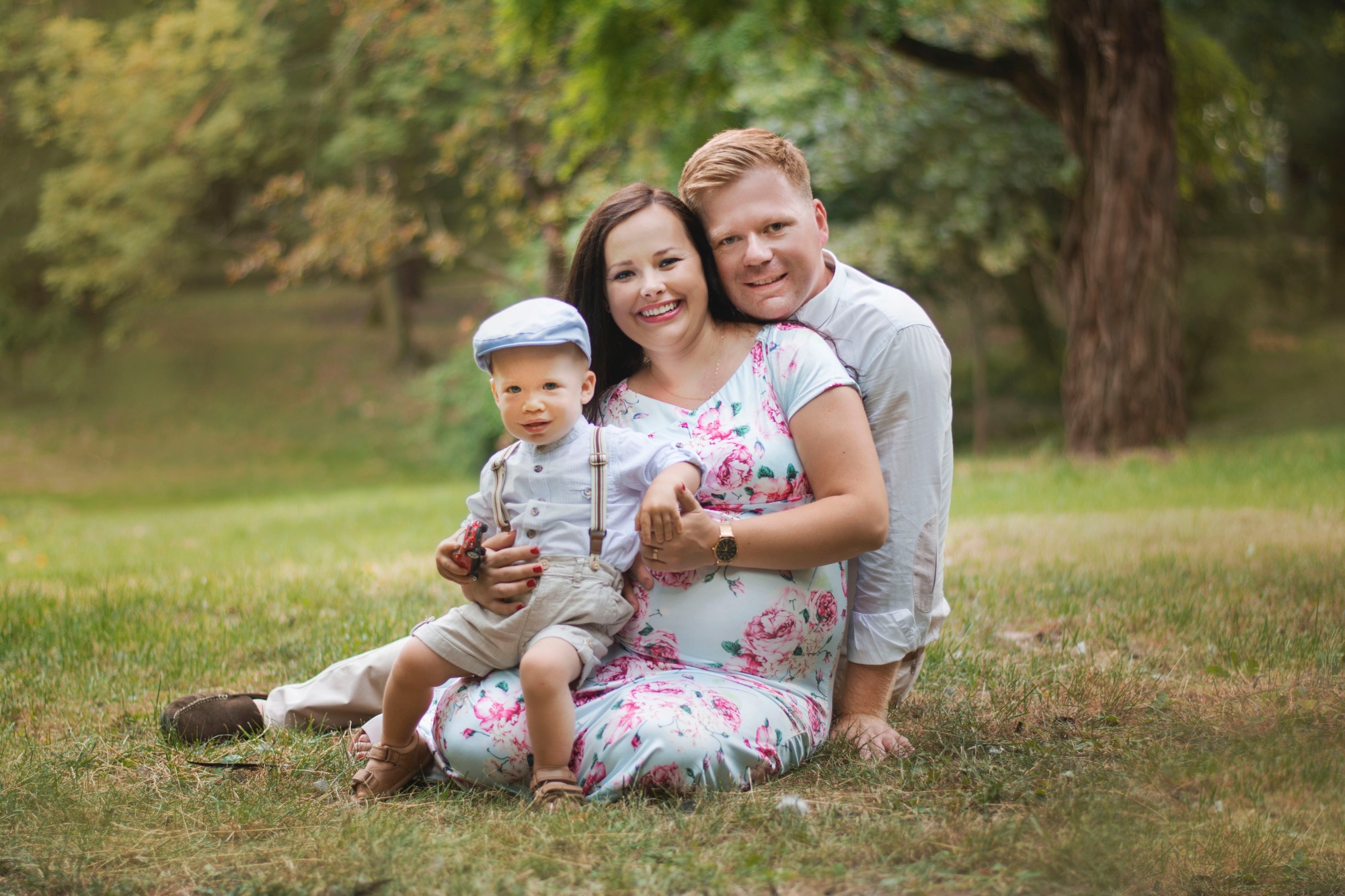 Szczęśliwa rodzina: ciężarna kobieta w kwiecistej sukience, mężczyzna w białej koszuli i mały chłopiec w retro stroju z szelkami i kaszkietem, siedzą na trawie w parku, chłopiec trzyma małą...