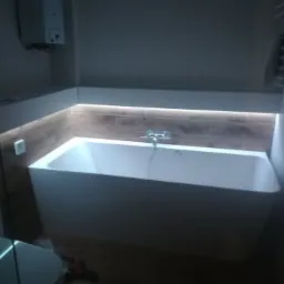 Nowoczesna, prostokątna wanna z podświetleniem LED w obudowie wykończonej płytkami imitującymi drewno, zamontowana w łazience w Gliwicach. Widoczny chromowany kran i fragment grzejnika.