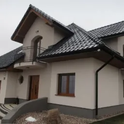 Nowoczesny dom z dwiema małymi balkonami z kutymi balustradami, grafitowym dachem i drewnianą stolarką okienną, otoczony szarą kostką brukową i zielenią.