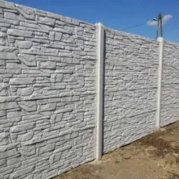 Długi, szary, betonowy mur z imitacją kamienia, podtrzymywany przez pionowe słupki, widoczny z boku pod lekkim kątem, na tle błękitnego nieba.