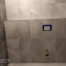 Ściana łazienki wyłożona szarymi płytkami imitującymi marmur, z widocznymi instalacjami hydraulicznymi i elektrycznymi przed montażem armatury.