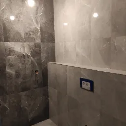 Wykończona łazienka z szarymi płytkami imitującymi marmur, widoczne przyłącza do toalety podtynkowej i gniazdko elektryczne bez osprzętu.