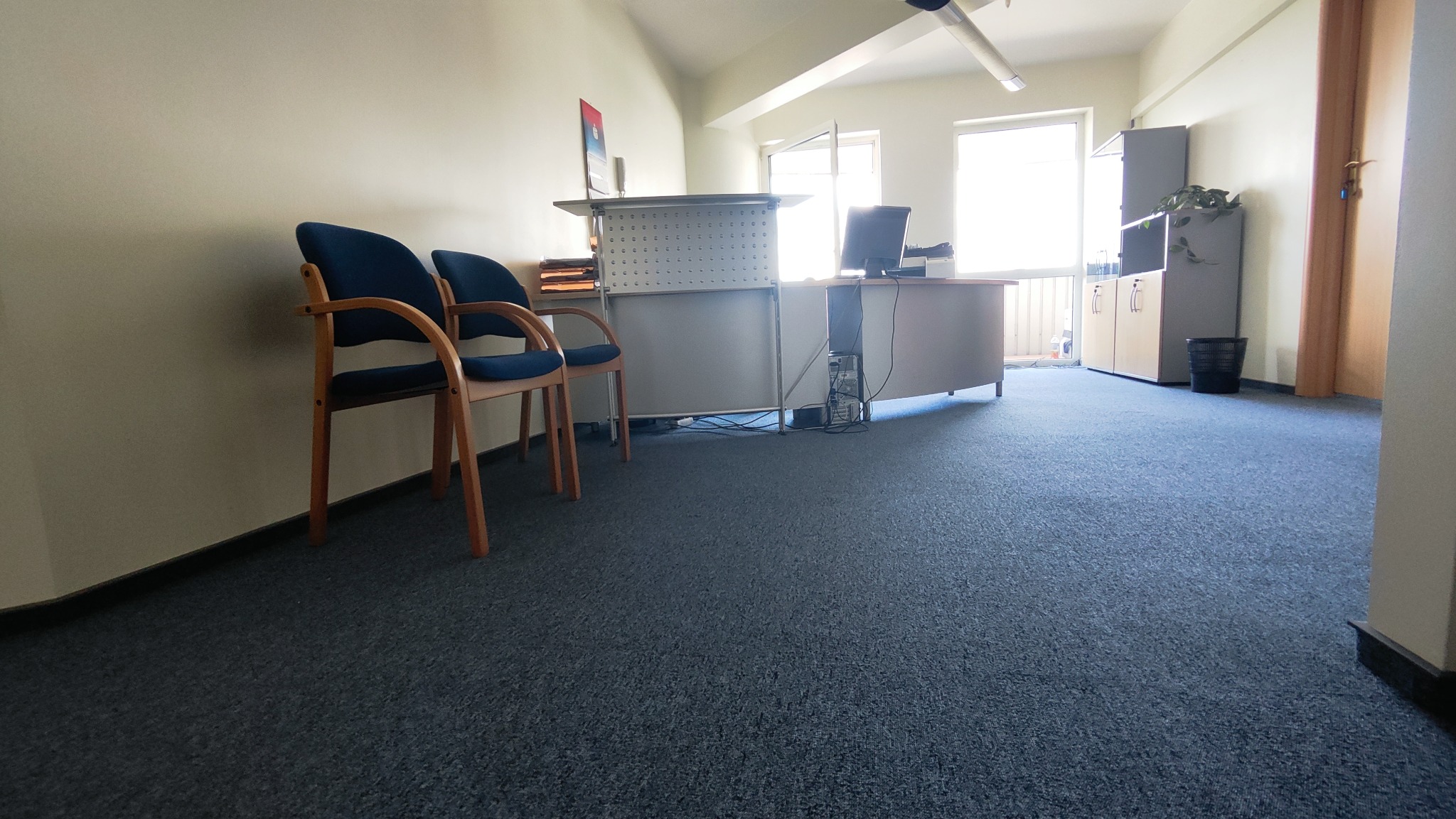 Poczekalnia z dwoma krzesłami z drewnianymi podłokietnikami i niebieskim obiciem, za nimi biurko recepcyjne, podłoga pokryta szarą wykładziną dywanową.