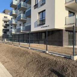 Nowoczesny budynek wielorodzinny z balkonami zabezpieczony ogrodzeniem panelowym w kolorze grafitowym, świeżo wyr&oacute;wnany teren przed budynkiem.