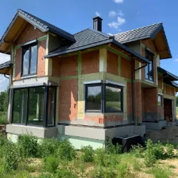 Budowa domu jednorodzinnego w stanie surowym, widoczne okna z roletami zewnętrznymi, ściany z cegły, dach z czarnej dachówki, zielone otoczenie.