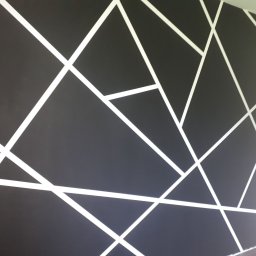 MK-WNĘTRZA - Czarna ściana z białymi, geometrycznymi wzorami tworzącymi abstrakcyjny design. Nowoczesne malowanie z użyciem taśmy malarskiej, widok z boku.