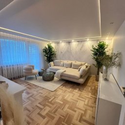 MK-WNĘTRZA - Elegancki salon z parkietem w jodełkę, beżową sofą, fotelami i roślinami. Sufit podwieszany z oświetleniem LED. Ściana z subtelnym wzorem liści. Nowoczesne wykończenie wnętrza.