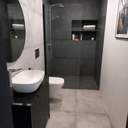 MK-WNĘTRZA - Nowoczesna łazienka z szarymi płytkami imitującymi beton, kabiną typu walk-in i owalnym lustrem. Umywalka nablatowa na czarnej szafce z ryflowanym frontem.