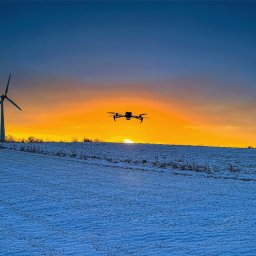 Four Winds - Dron w locie na tle zimowego krajobrazu o zachodzie słońca, w tle widoczna turbina wiatrowa i budynki z dymiącymi kominami.