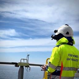 Four Winds - Operator drona w kasku i jaskrawej kurtce steruje dronem na tle morskiej farmy wiatrowej. Widoczny kontroler i turbina wiatrowa w oddali. Ujęcie z dołu.