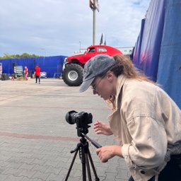 Four Winds - Kobieta ustawia aparat na statywie, przygotowując się do sesji zdjęciowej na zewnątrz. W tle czerwony monster truck i osoby w kostiumach, sugerujące plenerową produkcję.