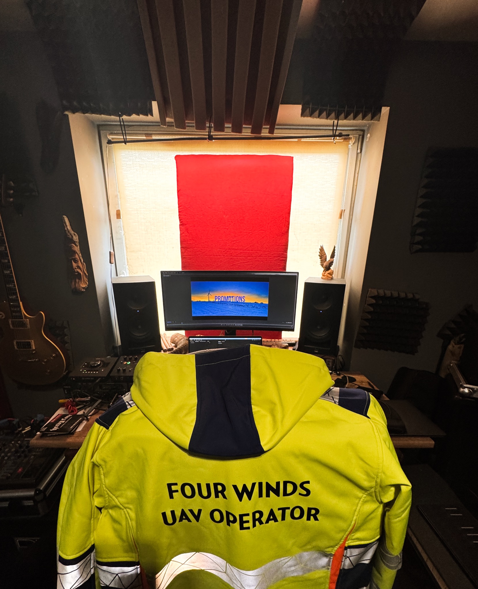 Studio montażu filmów z widokiem na monitor z napisem PROMOTIONS, żółtą kurtką UAV operatora i panelami akustycznymi na ścianach.