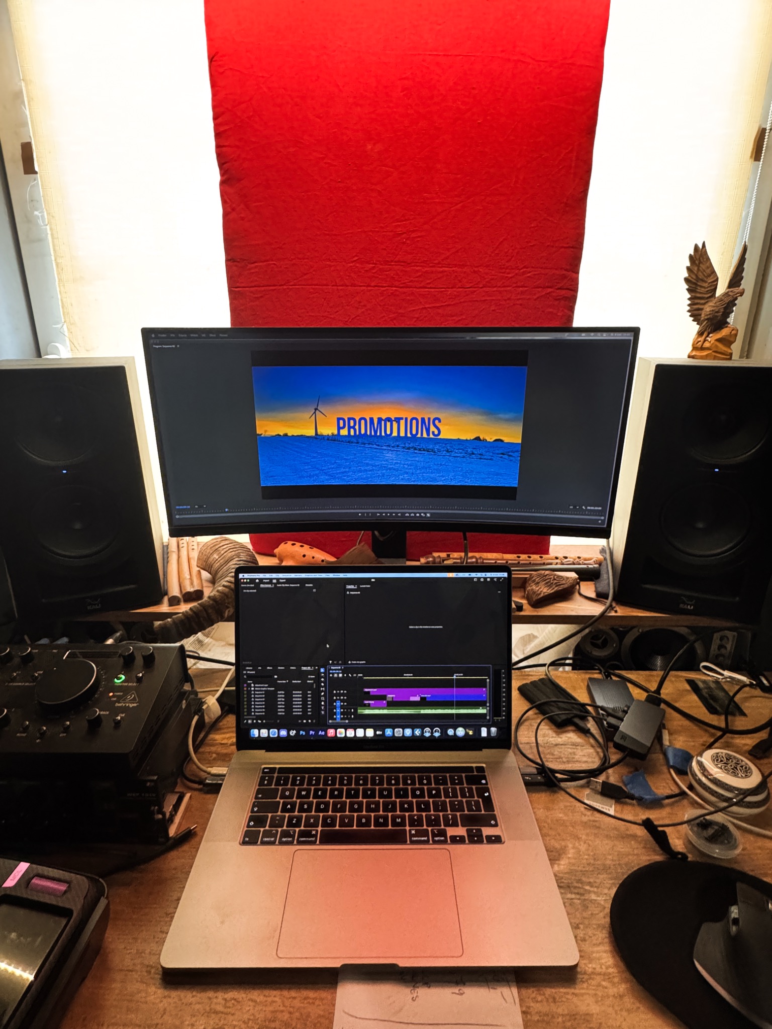 Stanowisko pracy z laptopem i monitorem wyświetlającym edytowany film z wiatrakiem na tle zachodzącego słońca, otoczone głośnikami i sprzętem audio, na drewnianym biurku z czerwonym tłem.