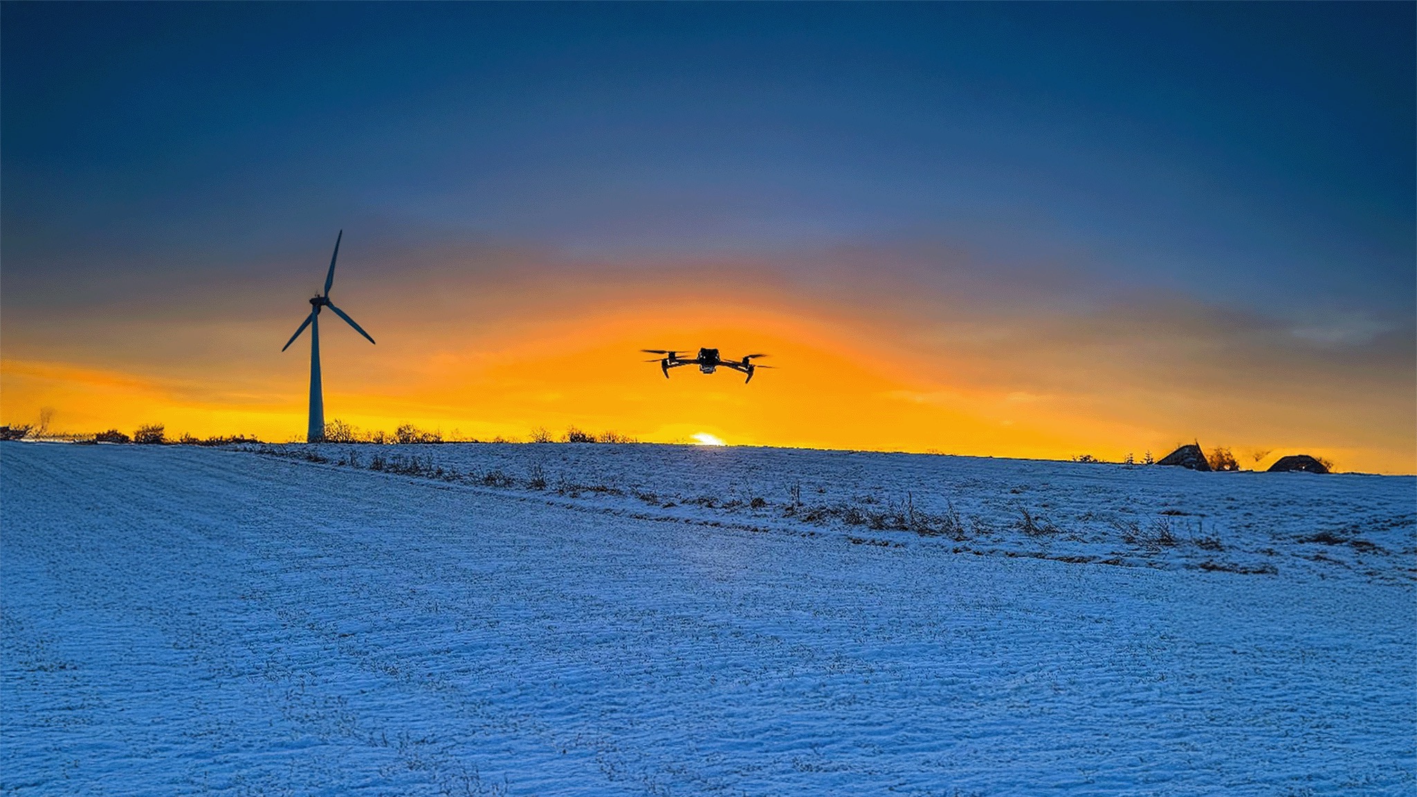 Dron w locie na tle zimowego krajobrazu o zachodzie słońca, w tle widoczna turbina wiatrowa i budynki z dymiącymi kominami.