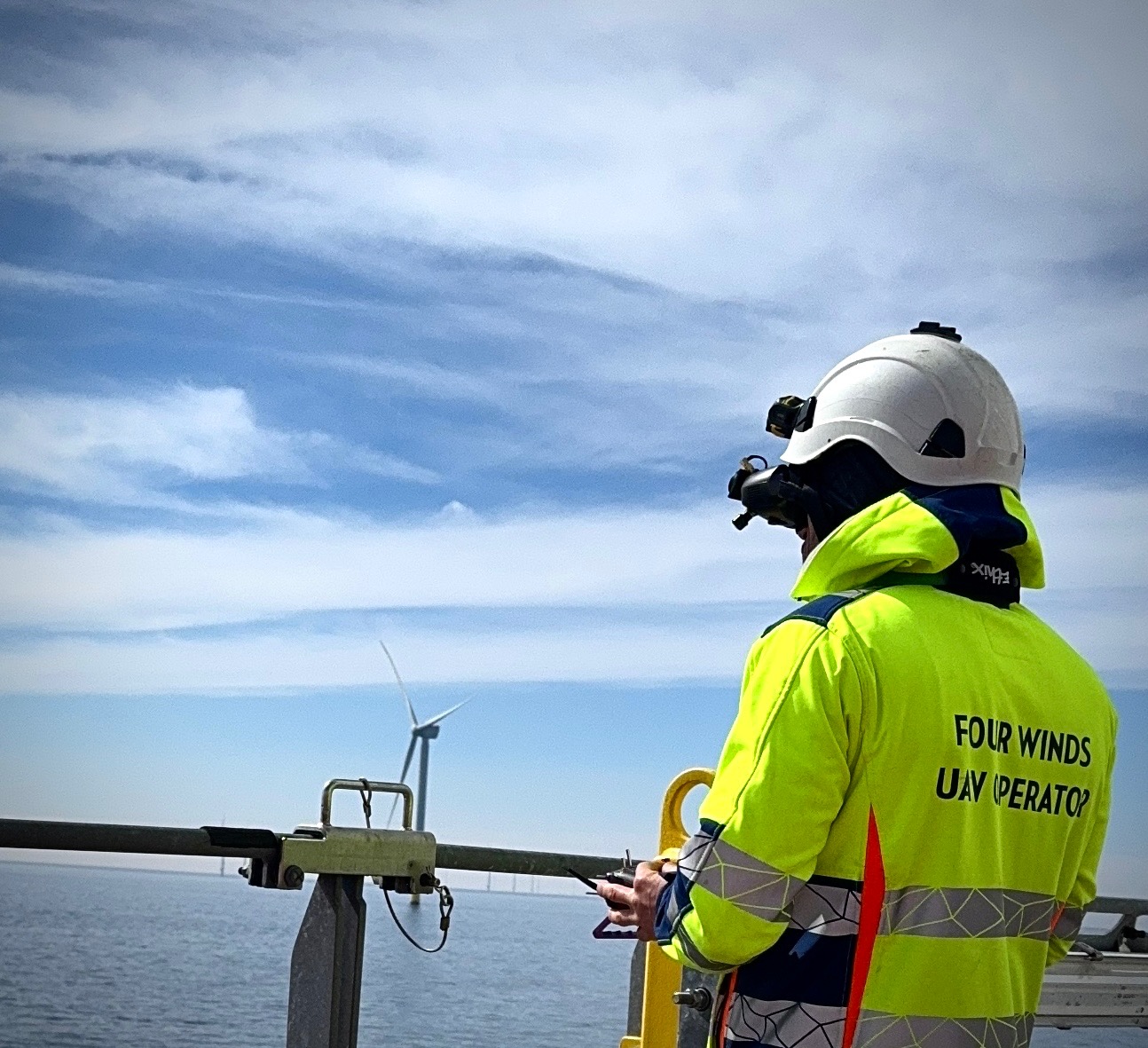 Operator drona w kasku i jaskrawej kurtce steruje dronem na tle morskiej farmy wiatrowej. Widoczny kontroler i turbina wiatrowa w oddali. Ujęcie z dołu.