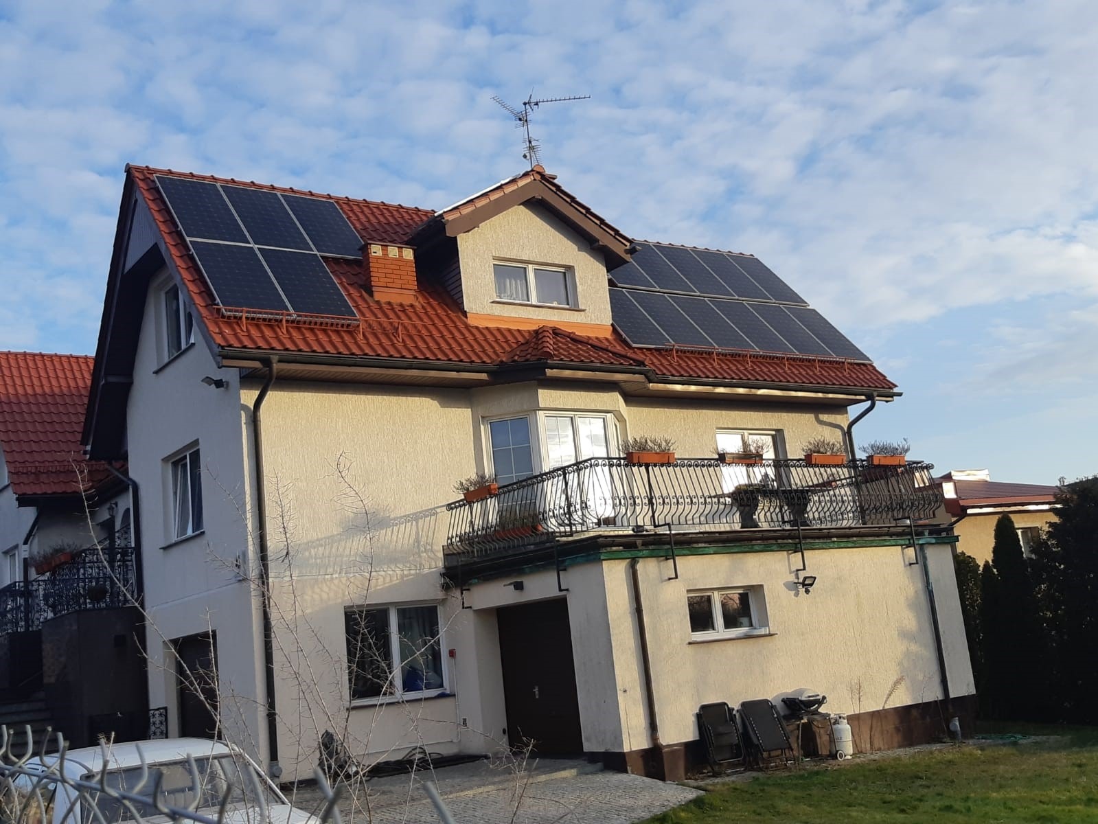 Dwukondygnacyjny dom jednorodzinny z panelami słonecznymi na dachu z czerwonej dachówki, widok z poziomu gruntu, błękitne niebo z chmurami w tle.