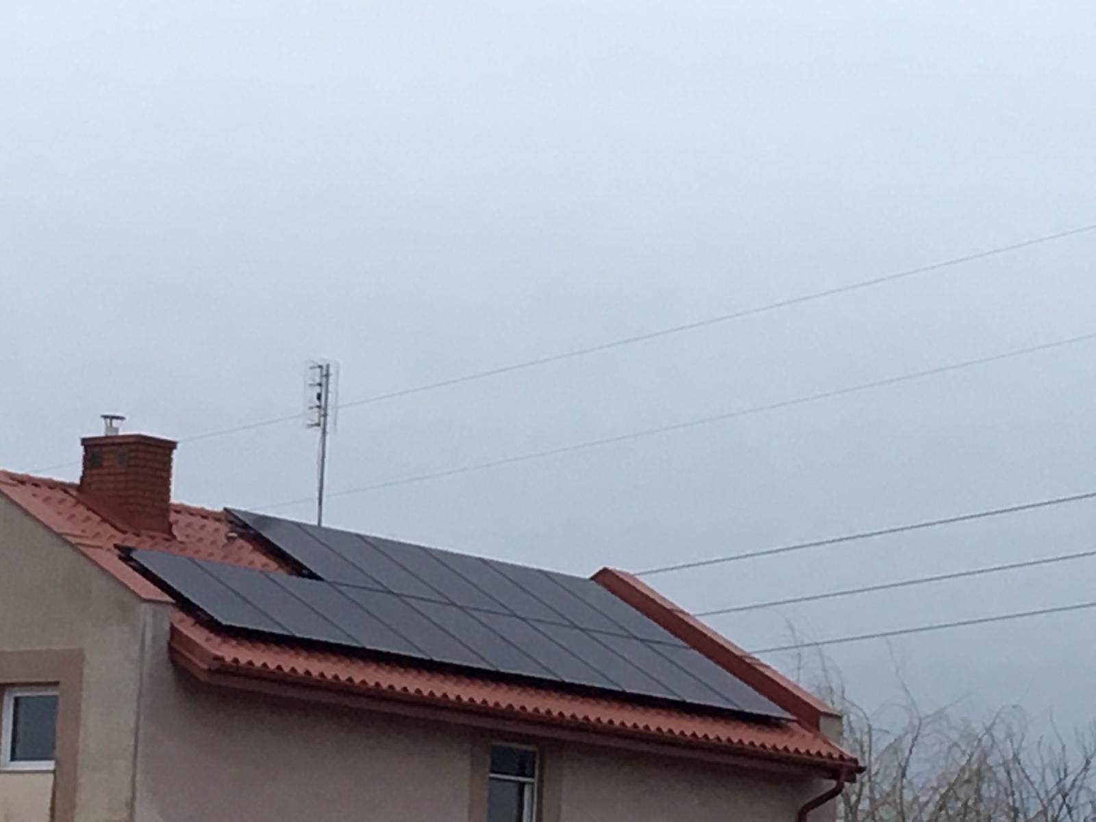 Część dachu domu jednorodzinnego z zainstalowanymi panelami słonecznymi w pochmurny dzień, widoczny komin i antena telewizyjna.