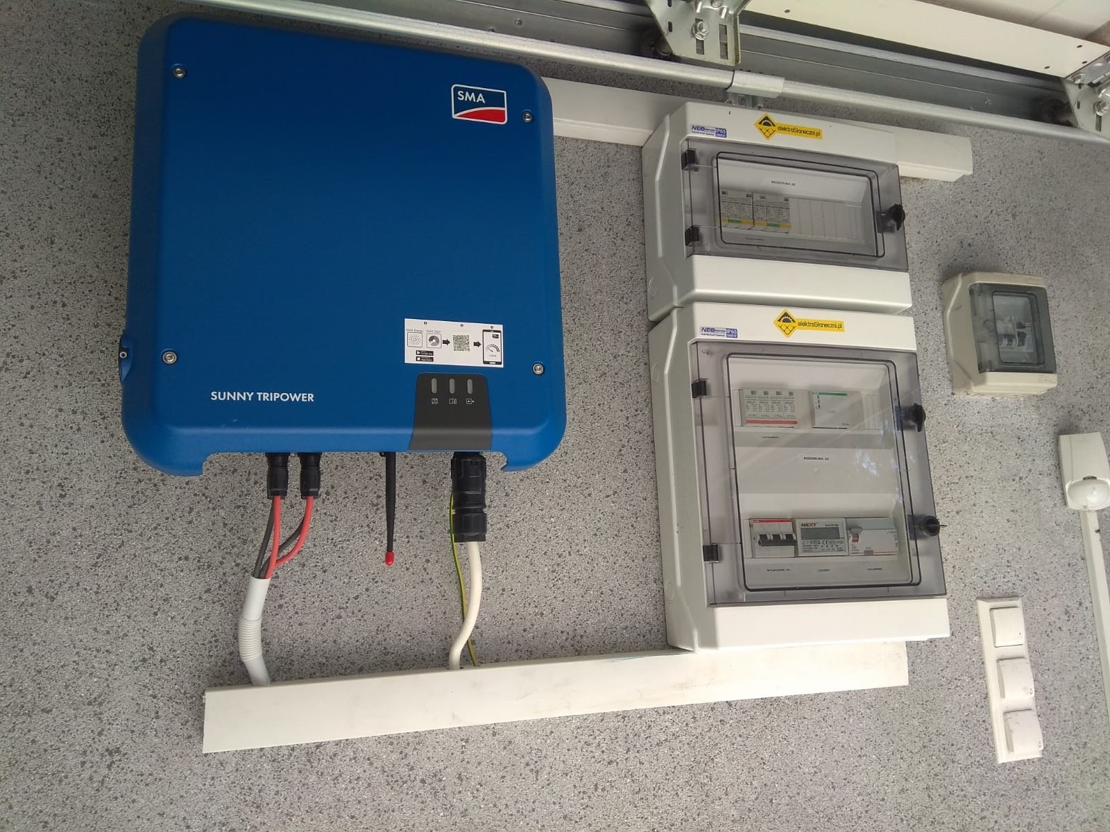 Niebieski inwerter SMA Sunny Tripower zamontowany na szarej ścianie obok dwóch białych skrzynek z zabezpieczeniami elektrycznymi.