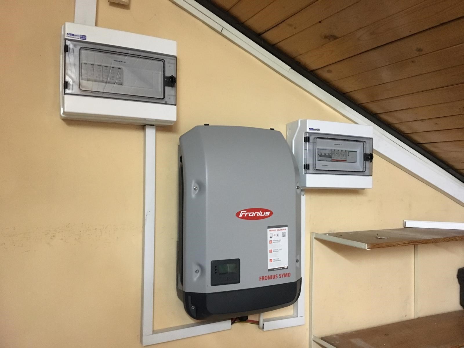 Szary falownik Fronius Symo zamontowany na żółtej ścianie z dwiema skrzynkami elektrycznymi, widoczne drewniane wykończenie sufitu.