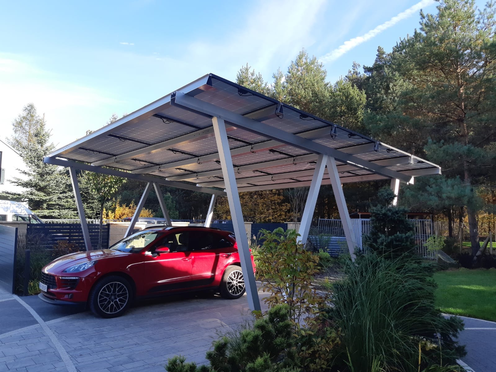 Czerwony samochód pod zadaszeniem z paneli słonecznych na tle zieleni i błękitnego nieba.