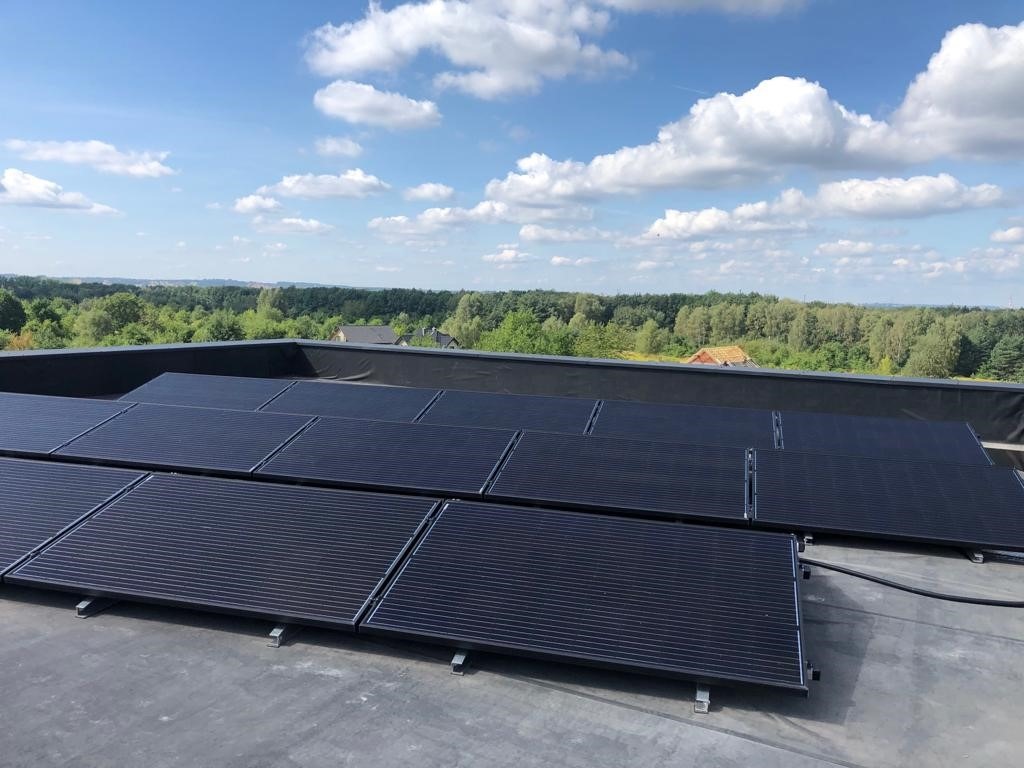 Panele słoneczne na płaskim dachu w Warszawie, widok na las i błękitne niebo z chmurami.
