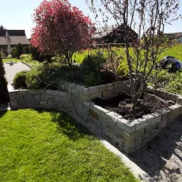 Kaskadowa rabata z kamienia naturalnego, obsadzona roślinami, w tym drzewkiem o purpurowych liściach, na tle zielonego trawnika i zabudowań.