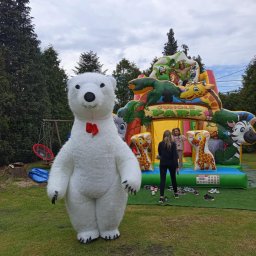 Zbigniew Paprocki - Duży kostium białego misia z czerwoną muchą na tle dmuchanego placu zabaw Jungle Park, z dwiema osobami i zdjętymi butami na sztucznej trawie.