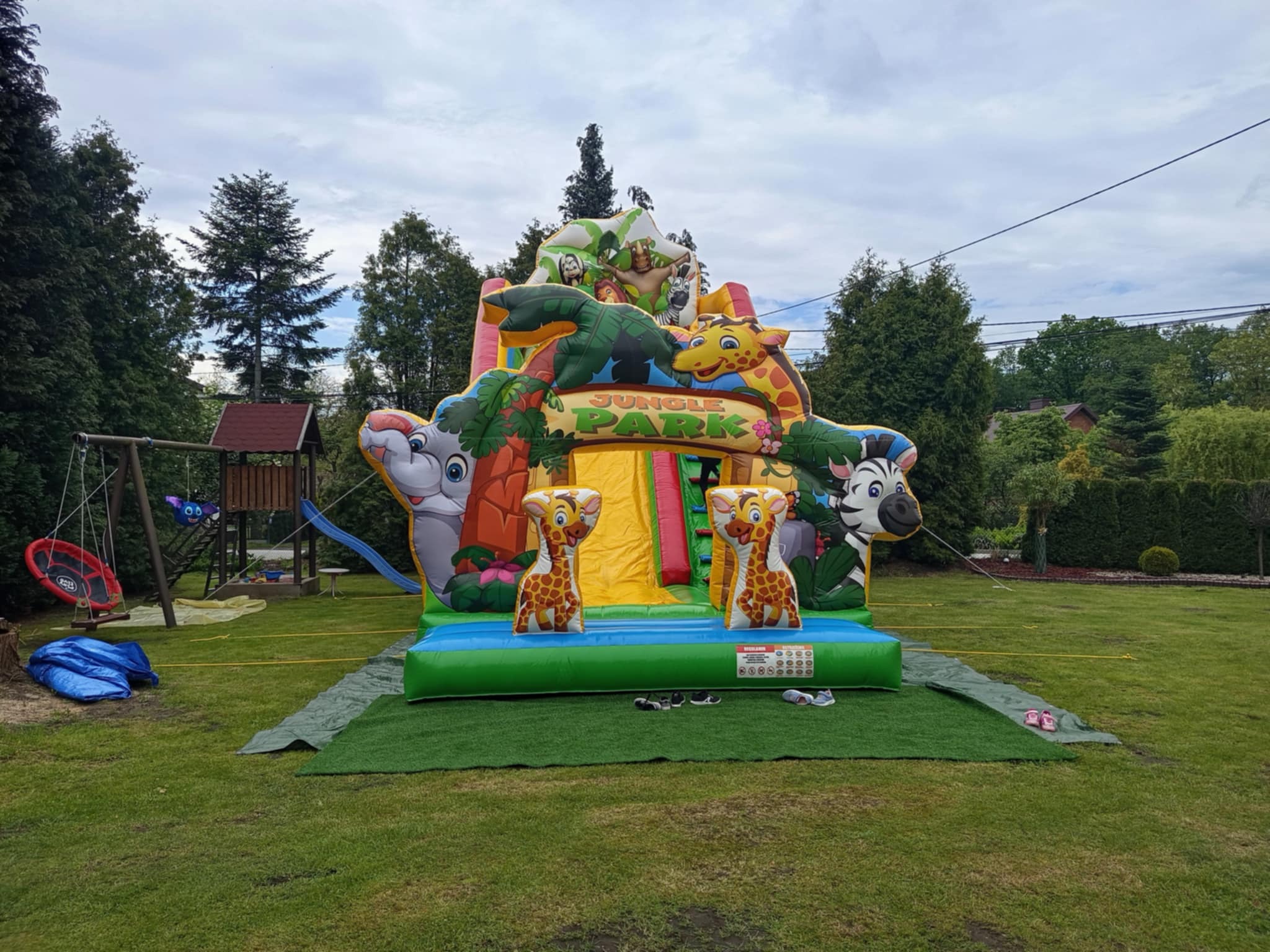 Dmuchany plac zabaw 'Jungle Park' z motywami zwierzęcymi na trawniku, otoczony zielenią. Widoczne drzewa i huśtawka w tle. Żywe kolory, pochmurne niebo.
