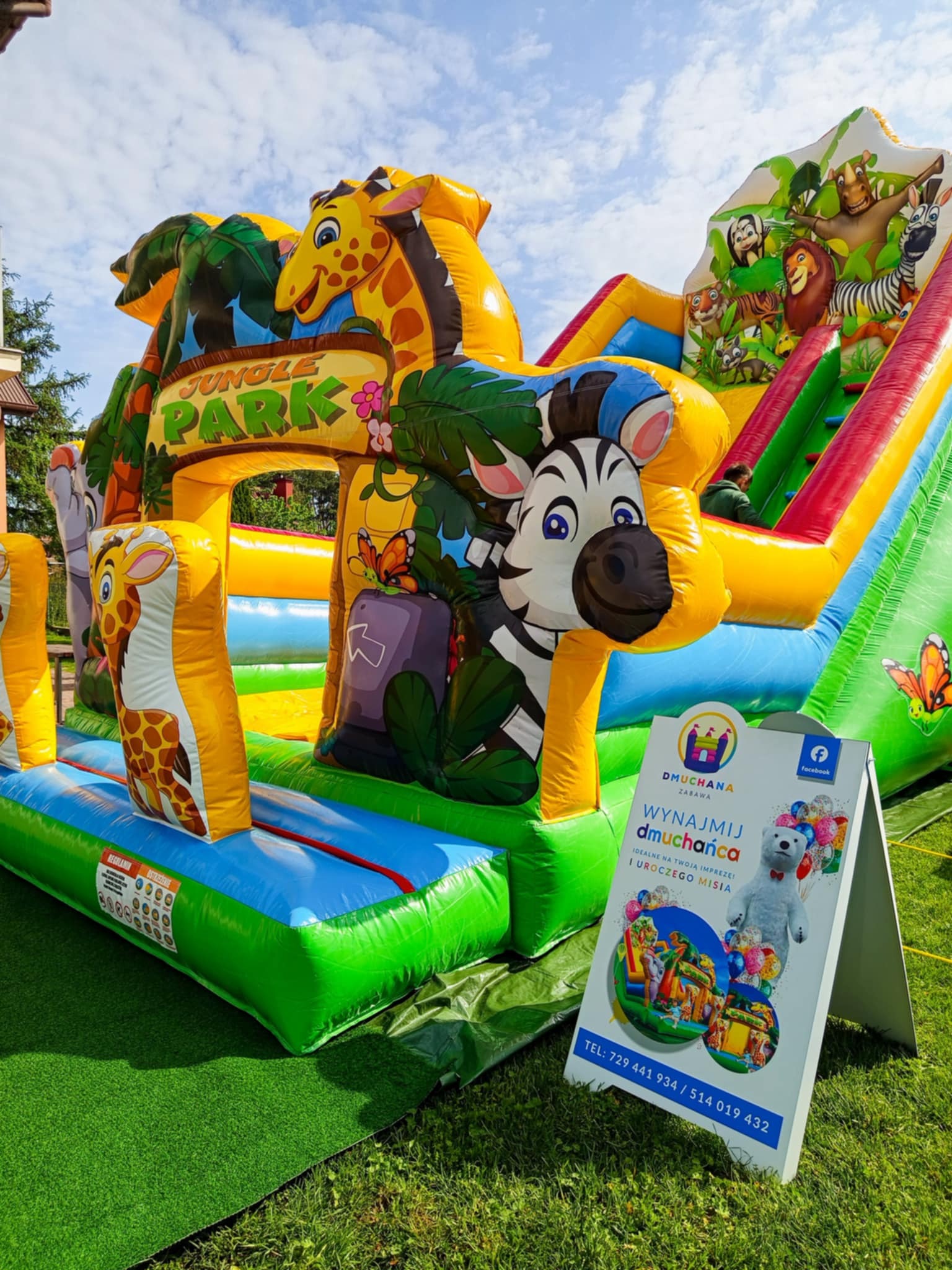 Kolorowy, dmuchany plac zabaw z motywem dżungli, żyrafa i zebra, zjeżdżalnia, na trawie, obok reklama firmy 'Dmuchana Zabawa' z misiem i numerem telefonu.
