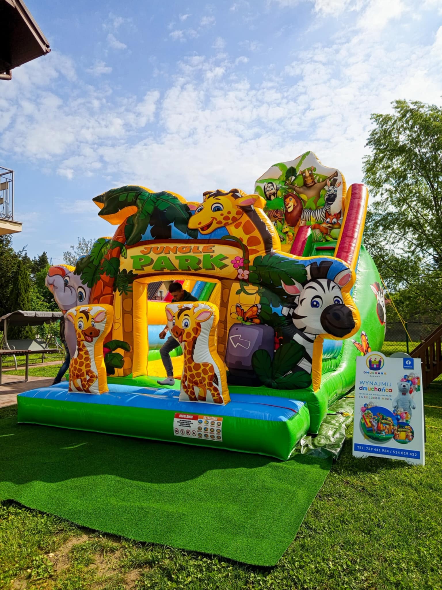 Kolorowy, dmuchany plac zabaw 'Jungle Park' z motywami zwierzęcymi, ustawiony na zielonej trawie. Widoczna tablica reklamowa z ofertą wynajmu dmuchańców.
