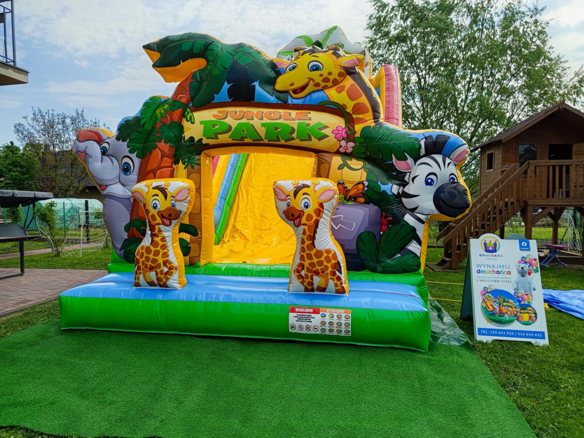 Dmuchany zamek 'Jungle Park' z żyrafami, słoniem i zebrą. Kolorowa atrakcja na trawie, obok tablica z ofertą wynajmu dmuchańców na imprezy.