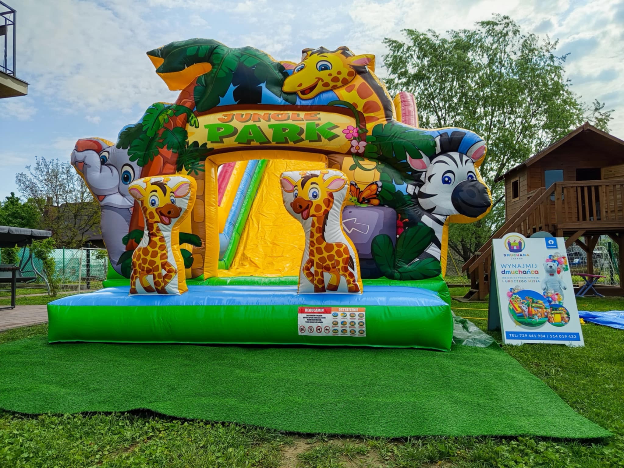 Kolorowy, dmuchany zamek w stylu dżungli z motywami zwierzęcymi (żyrafy, zebra, słoń) na zielonej trawie. Widoczny baner reklamowy firmy oferującej wynajem.