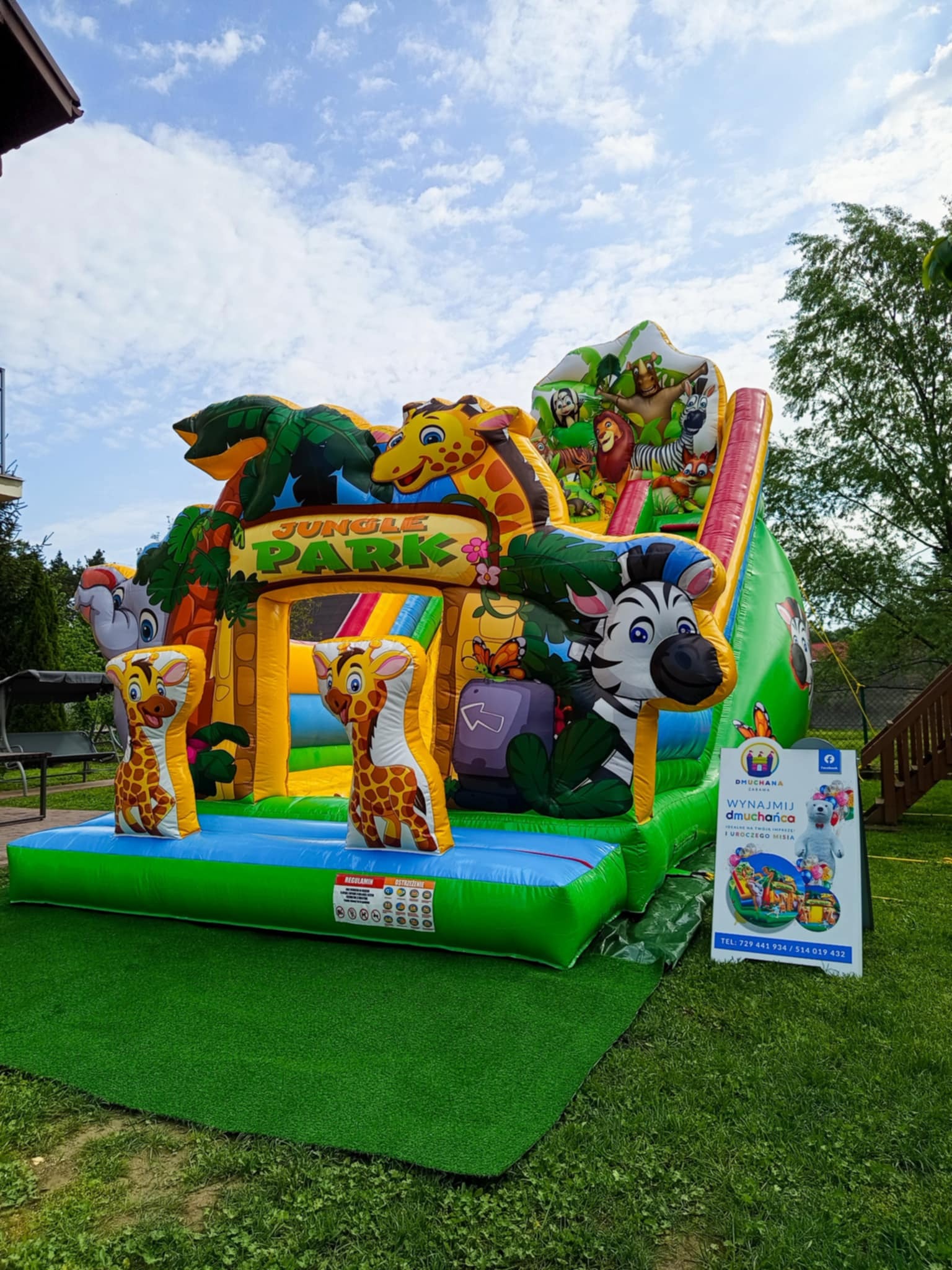 Kolorowy, dmuchany plac zabaw w stylu dżungli z żyrafami, zebrą i zjeżdżalnią, ustawiony na trawie. Obok tablica reklamowa z napisem 'Wynajmij dmuchańca'.