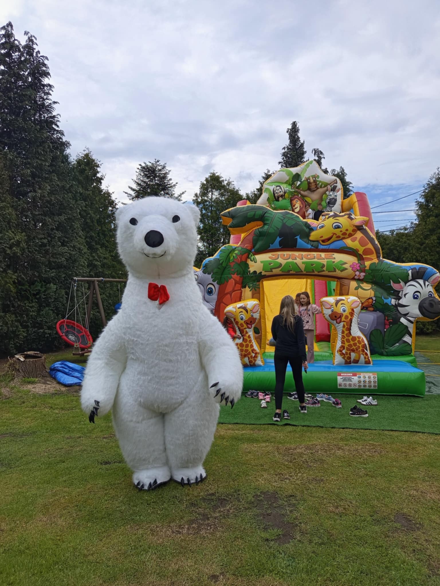 Duży kostium białego misia z czerwoną muchą na tle dmuchanego placu zabaw Jungle Park, z dwiema osobami i zdjętymi butami na sztucznej trawie.