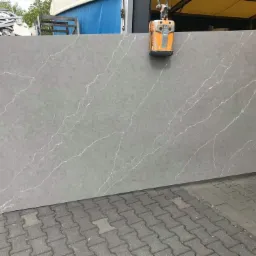 Szary blat z konglomeratu kwarcowego z białymi żyłkami, podnoszony przez pomarańczowy chwytak próżniowy, na tle stosu płyt i kostki brukowej.