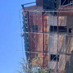 Wznoszona konstrukcja dachu z cegły ceramicznej i metalowych elementów więźby, widoczne rusztowania i kwitnące drzewo na pierwszym planie.