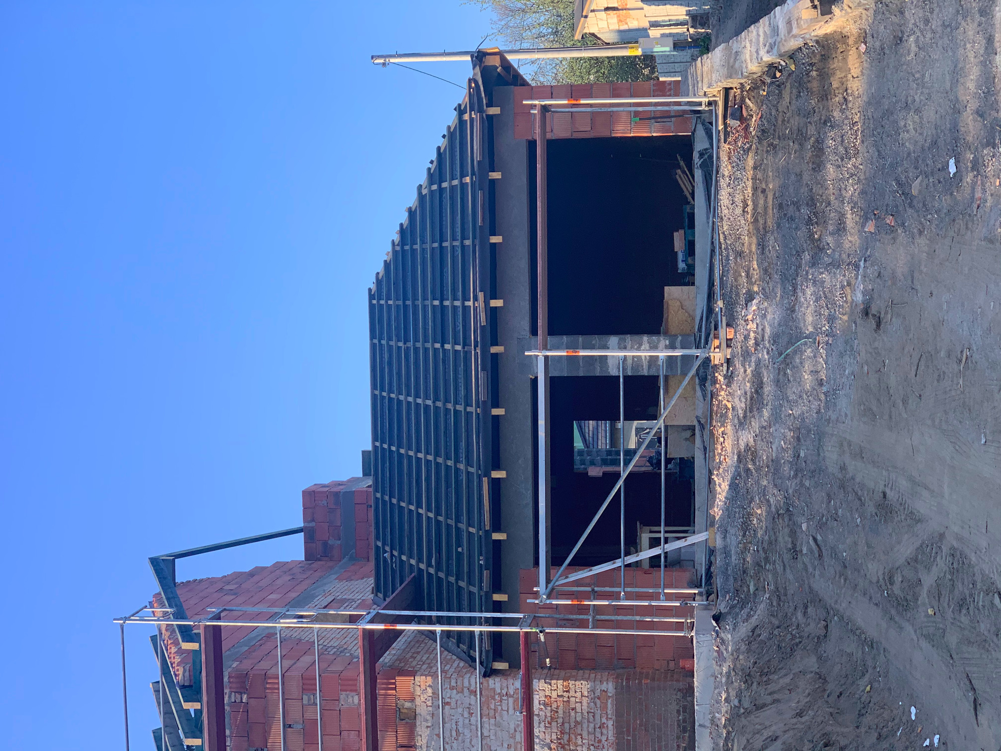 Wznoszenie konstrukcji dachu z widoczną więźbą i deskowaniem, ceglany budynek w trakcie budowy, rusztowania w tle, słoneczny dzień.