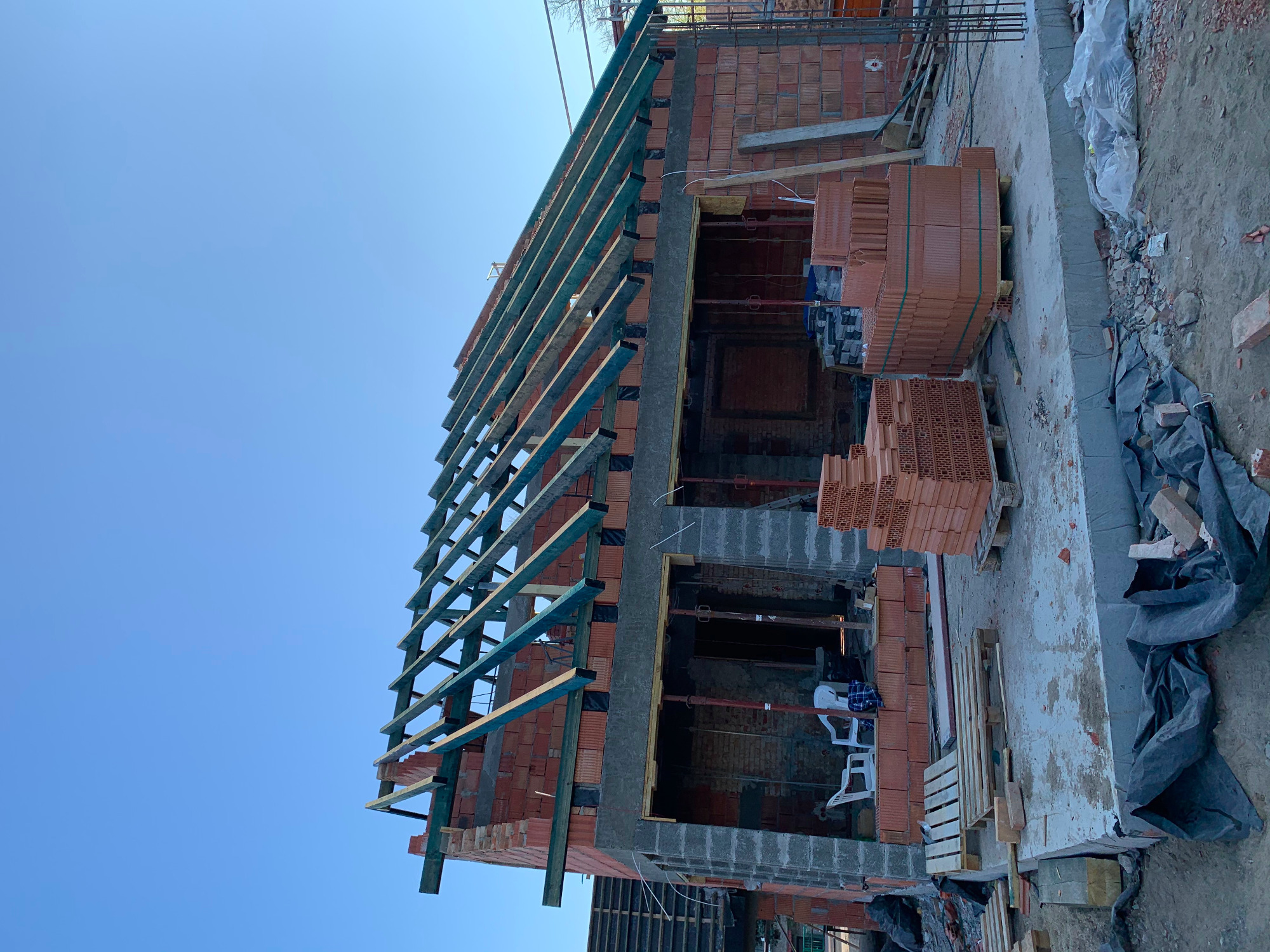 Wznoszona dwukondygnacyjna konstrukcja budynku z czerwonej cegły z widoczną drewnianą konstrukcją dachu i paletami cegieł na placu budowy, fotografowana z dołu pod kątem w słoneczny dzień.