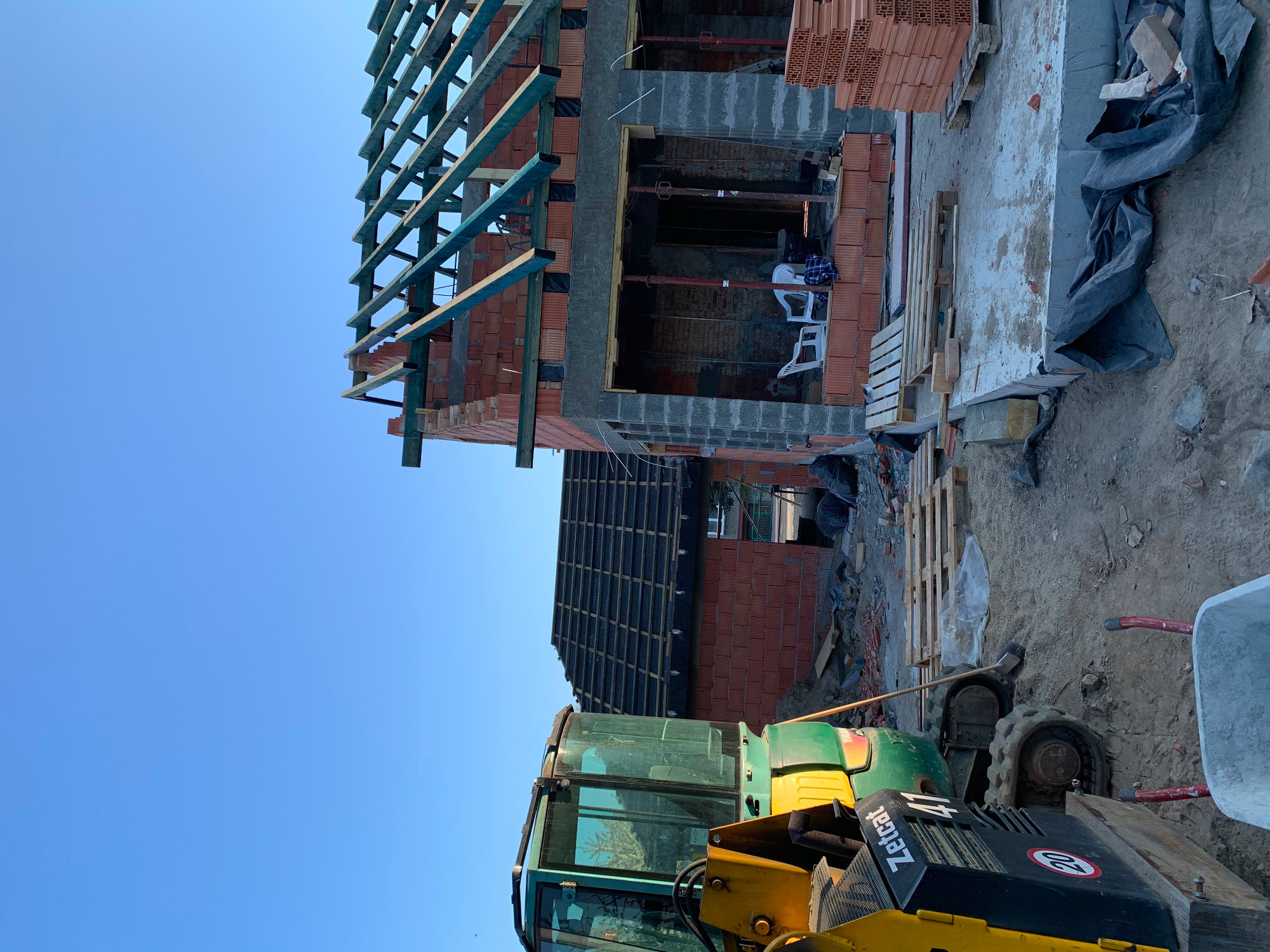 Budowa domu w trakcie: widoczna konstrukcja więźby dachowej, ściany z czerwonej cegły oraz maszyna budowlana na pierwszym planie.