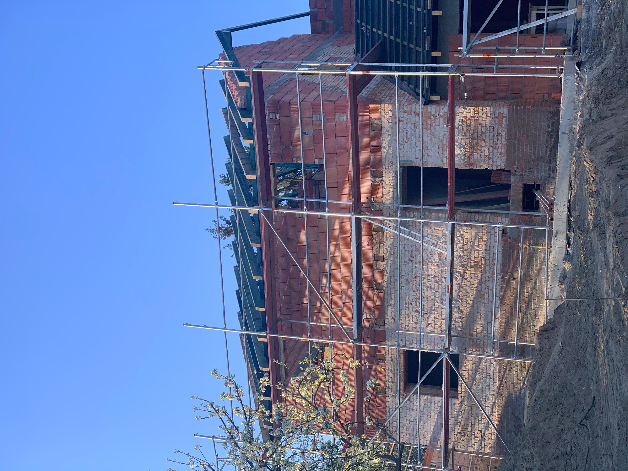 Wznoszona konstrukcja dachu z cegły ceramicznej i metalowych elementów więźby, widoczne rusztowania i kwitnące drzewo na pierwszym planie.