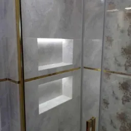 Wykończona łazienka z szarymi płytkami imitującymi marmur, wnękami z podświetleniem LED i złotymi akcentami na kabinie prysznicowej.