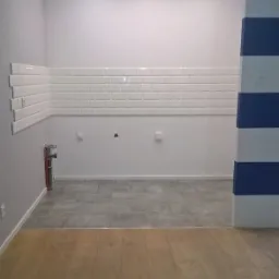 Wnętrze w trakcie prac: częściowo ułożone białe płytki typu metro na ścianie, szara podłoga z płytek, drewniana podłoga w sąsiednim pomieszczeniu, kolumna pomalowana w biało-niebieskie pasy.