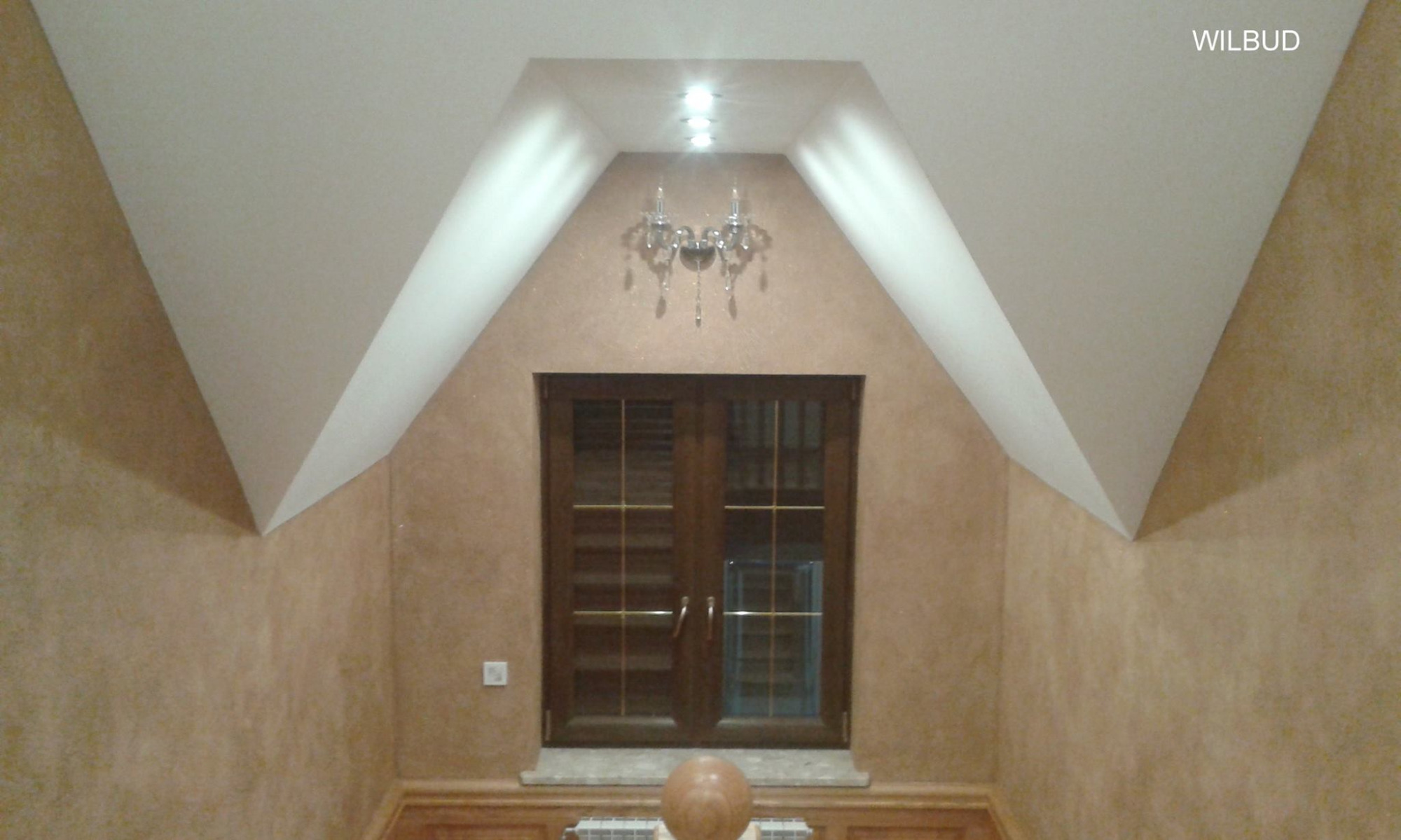 Pokój na poddaszu z oknem dwuskrzydłowym, ściany w odcieniach beżu z dekoracyjną fakturą, biały sufit ze skosami i żyrandolem